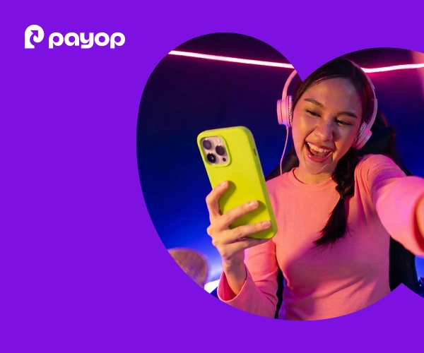 A woman joyfully holds a green phone, wearing pink headphones and a top, smiling during a video call in a neon-lit room. The background features a purple overlay and the Payop logo.