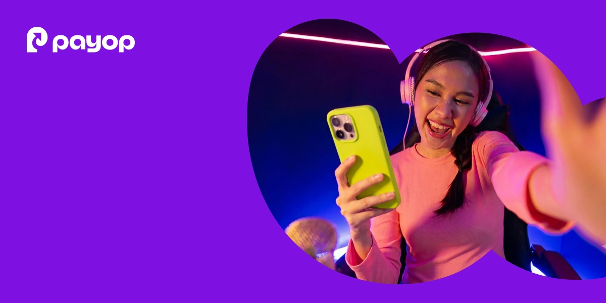 A woman joyfully holds a green phone, wearing pink headphones and a top, smiling during a video call in a neon-lit room. The background features a purple overlay and the Payop logo.